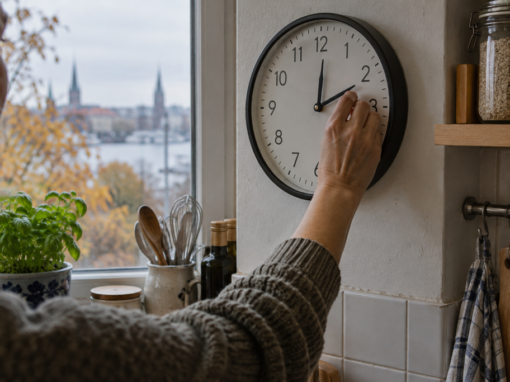 Eine Person verstellt die Zeiger einer runden Wanduhr in einer Küche. Durch das Fenster sind herbstliche Bäume und eine Stadtsilhouette zu sehen.